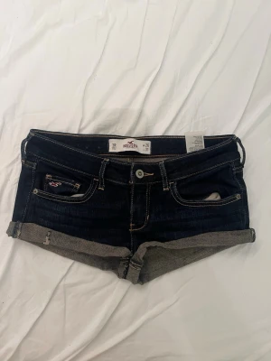 Mörkblå denimshorts från hollister  - Säljer ett par mörkblå denimshorts med låg midja och uppvikta benslut. Shortsen har slitna detaljer på bakfickorna och kontrastsömmar. Klassisk femficksmodell med knapp och dragkedja framtill. Midjemåttet rakt över : 39cm, längden på shortsen : 26cm