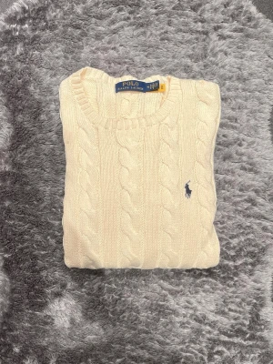 Kabelstickad tröja från Polo Ralph Lauren - Kabelstickad cremevit tröja från Polo Ralph Lauren med rund halsringning och blå broderad logga på bröstet. Tröjan har långa ärmar och ribbade muddar vid ärmslut och nederkant. Perfekt för lager på lager och klassisk stil.