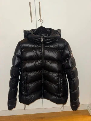 Colmar jacka dam - Snygg svart dunjacka från Colmar med glansig finish och stor vadderad huva. Jackan har breda kanaler, två sidofickor med dragkedja och en Colmar-logga på ärmen. Perfekt för kalla vinterdagar och riktigt varm tack vare dunfyllningen.
