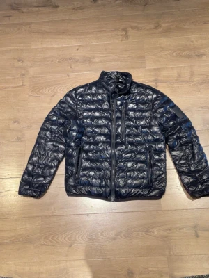 Svart pufferjacka från Peak Performance - Snygg svart pufferjacka från Peak Performance med hög krage och tre dragkedjeförsedda fickor framtill. Jackan har ett diskret logomärke på ärmen och quiltad design som ger en modern look. Perfekt för kalla dagar och enkel att matcha med olika outfits.