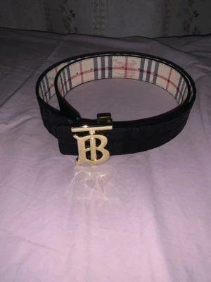 Svart Burberry bälte med guldspänne - Snyggt svart bälte från Burberry med klassiskt rutigt innerfoder och ett stort guldigt TB-spänne framtill. Bältet har diskret präglad logga på utsidan och är perfekt för att lyfta vilken outfit som helst.