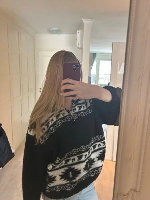 Svart och vit mönstrad stickad tröja - En oversized svart stickad tröja med vit geometriskt mönster över bröst och mage. Tröjan har en hög krage med dragkedja och långa ärmar. Perfekt för att hålla sig varm och snygg under kalla dagar. Den här tröjan har inte använts mycket.