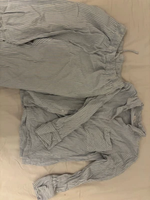 Randig pyjamas i bomull - Klassisk tvådelad pyjamas med ljusblå och vita smala ränder. Skjortan har knappar framtill och en bröstficka, byxorna har resår och dragsko i midjan. Tillverkad i mjuk bomull för extra komfort, perfekt för mysiga kvällar hemma.