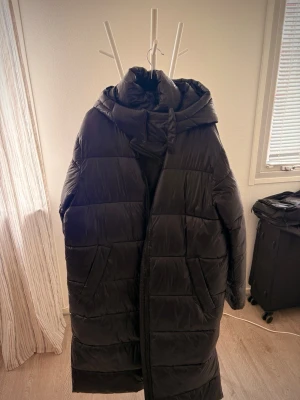 Svart lång pufferjacka med huva - Säljer en svart lång pufferjacka från 157 med stor vadderad huva och dragkedja framtill. Jackan har två stora fickor på sidorna och quiltad design. Perfekt för kalla vinterdagar och riktigt skön att bära.