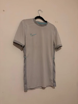 Ljusgrå sporttröja från Nike - Snygg ljusgrå sporttröja från Nike med turkos logga på bröstet. Tröjan har korta ärmar och meshpaneler på sidorna för extra ventilation. Tillverkad i ett lätt och snabbtorkande funktionsmaterial som passar perfekt till träning.