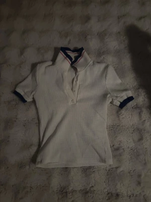 Vit henley med blå detaljer - Vit ribbad henley med korta ärmar och blå muddar. Kragen har en sportig look med rosa och blå rand. Knappar framtill för en klassisk touch. Perfekt för en avslappnad och stilren outfit.