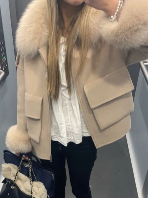 Beige kappa med fukpäls från FUR - Trendig beige kappa från FUR Stockholm med stora fickor, dragkedja och fluffiga detaljer i päls vid kragen och ärmslut. Kortare modell som ger en cool och lyxig vibe. Perfekt för dig som vill sticka ut med en statement-jacka i höst och vinter.