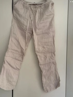 Beiga vida linnebyxor med resår - Säljer ett par beiga linnebyxor med avslappnad passform och resår i midjan. Byxorna är raka i modellen och har en luftig känsla, perfekta för varma dagar. Tillverkade i linne för en skön och sval stil.