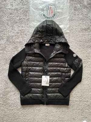 Svart pufferjacka från Moncler - Svart pufferjacka från Moncler med stickade ärmar och vadderad kropp. Jackan har huva, dragkedja framtill och Moncler-logga på vänster ärm. Materialmix av glansigt nylon och stickat tyg ger en cool kontrast. Perfekt för kalla dagar. Den kan passa M beror på längd och vikt hör av er vid intresse