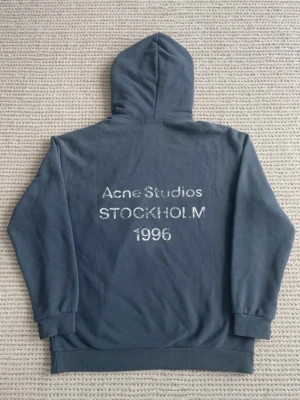 Blå hoodie från Acne Studios - Snygg blå hoodie från Acne Studios med tryck på ryggen där det står 'Acne Studios STOCKHOLM 1996' i vitt. Hoodien har huva och ribbade muddar vid ärmslut och nederkant. Perfekt för en avslappnad och trendig stil.