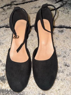 Svarta klackskor med ankelrem - Stilrena svarta pumps med rund tå och bred klack. Skorna har en elegant ankelrem med spänne och är tillverkade i ett mockaliknande material. Använda 1 gång 