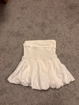 Sommartopp - Supersöt vit bandeau tubtopp från Gina Tricot med volang nertill. Toppen är axelbandslös och har en flowy, luftig passform nedtill. Perfekt att styla med jeans eller kjol för en trendig look.