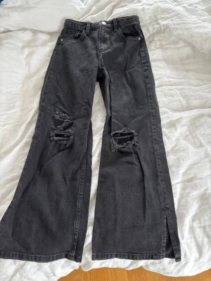 Svarta vida jeans med slitningar - Säljer ett par svarta jeans med breda ben och slitningar på båda knäna. Jeansen har hög midja, fem fickor och slitsar längst ner på benen. Materialet är denim i bomull och de har en cool, avslappnad vibe🩶😍