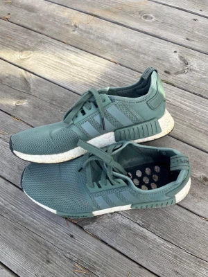 Adidas NMD R1 gröna sneakers - Snygga Adidas NMD R1 sneakers i en dämpad grön färg med klassiska tre ränder på sidan. Ovandelen är i meshmaterial för bra andningsförmåga och sulan är vit med Boost-dämpning. Skorna har lågt skaft och snörning framtill.