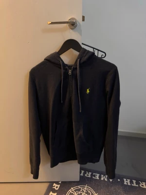 Mörkblå hoodie från Polo Ralph Lauren - Mörkblå hoodie med dragkedja från Polo Ralph Lauren. Klassisk design med huva och dragsnören, känguruficka framtill och den ikoniska gula loggan broderad på bröstet. Mjuk bomullsblandning som känns skön mot huden. Perfekt för en avslappnad och stilren look.det är skön vintage look och passar som 165-175