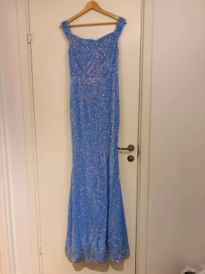 Blå glittrig långklänning med slits - Superfin blå långklänning täckt av glittrande paljetter. Klänningen har breda axelband, figurnära passform och en snygg slits framtill. Perfekt för bal eller festkvällar när du vill sticka ut. Använd 1 gång