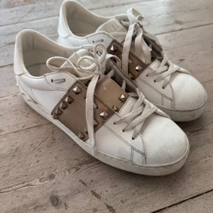 Valentino Rockstud vita sneakers - Snygga vita Valentino Rockstud sneakers i skinn med ikoniska nitar i silver på beige band över vristen. Klassisk rund tå, platt sula och snörning framtill. Perfekta för dig som vill ha en lyxig och edgy touch till din outfit. Vid en snabb affär kan priset rabatteras 