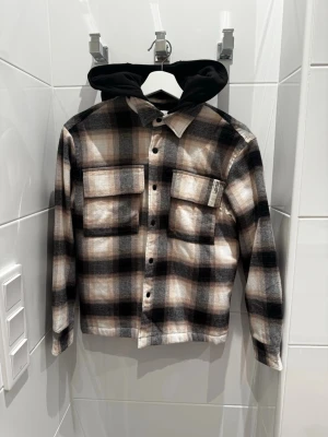Rutig overshirt med huva från Zara - Snygg overshirt från Zara i svart, beige och grått rutmönster med svarta knappar och två stora bröstfickor. Har en svart huva i sweatshirtmaterial som ger en cool lager-på-lager-look. Perfekt för att lyfta din outfit med streetkänsla. OBS! Luvan går att ta av, den sitter fast med knappar.