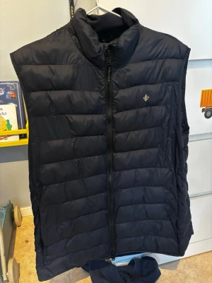 Svart dunväst från Morris - Snygg svart dunväst från Morris med quiltad design och dragkedja framtill. Västen har ståkrage och en liten broderad logga på bröstet. Perfekt att slänga över en hoodie eller tröja för extra värme. Klassisk och stilren look som funkar till det mesta.