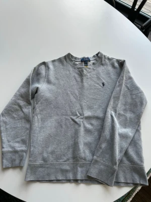 Grå sweatshirt från Polo Ralph Lauren stl 14-16 år - Klassisk grå sweatshirt från Polo Ralph Lauren med rund halsringning och broderad marinblå logga på bröstet. Tröjan har ribbade muddar vid ärmslut och nederkant. Perfekt för en avslappnad och stilren look. Obs liten i storleken. 