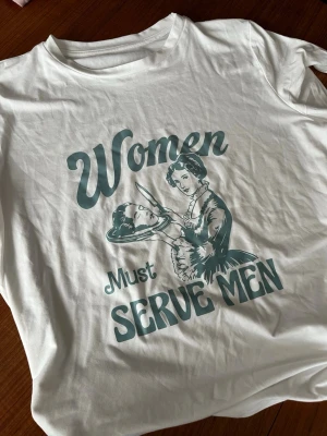 Vit t-shirt med retrotryck - Vit t-shirt med rund hals och tryck i retrostil som visar en kvinna med en maträtt och texten 'Women Must Serve Men'. T-shirten har korta ärmar och är gjord i mjukt bomullsmaterial. Perfekt för dig som gillar statement-plagg.