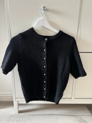 Svart kortärmad kofta med knappar - Stilren svart kofta med korta ärmar och rund halsringning. Koftan har dekorativa knappar framtill och är stickad i ett mjukt material. Perfekt att bära över ett linne eller t-shirt för en enkel men snygg look. Använd en gång och är som ny💕