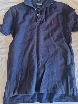 Marinblå pikétröja från Polo Ralph Lauren - Snygg marinblå pikétröja från Polo Ralph Lauren med klassisk krage och två knappar. Tröjan har det ikoniska röda broderade logotypen på bröstet och är tillverkad i bomull. Perfekt för en avslappnad och stilren look. Pris kan diskuteras. 