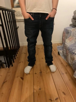 Mörkblå slim fit jeans herr från Levis - Säljer ett par mörkblå slim fit jeans med klassisk femficksdesign och normalhög midja. Jeansen har snygga slitningar och kontrastsömmar, perfekta till en avslappnad stil. Materialet är troligtvis bomull med lite stretch för extra komfort.