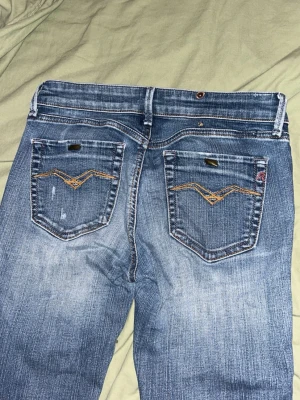 Blå bootcut jeans med broderi - Snygga blå bootcut jeans med slitningar och broderade detaljer på bakfickorna. Jeansen har en klassisk femficksmodell och kontrastsömmar. Perfekta till sneakers eller boots för en avslappnad look.