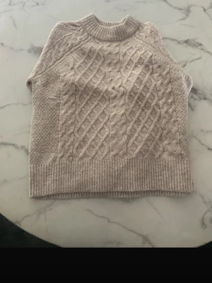 Beige kabelstickad tröja - Säljer en beige kabelstickad tröja med grovt mönster framtill och ribbade muddar vid ärmslut och nederkant. Tröjan har rund halsringning och är gjord i ett mjukt, tjockt material som känns riktigt cozy.