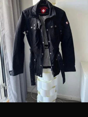 Svart trenchcoat från Wellensteyn - Snygg svart trenchcoat från Wellensteyn med bälte i midjan, dubbla bröstfickor med knappar och detaljer i metall. Jackan har klassisk krage, långa ärmar och logotyp på ärmen. Perfekt för dig som vill ha en stilren och funktionell jacka.