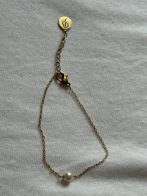 Edblad Guldfärgat armband med pärla - Snyggt och minimalistiskt armband i guldfärgad metall med en ensam vit pärla som detalj. Kedjan är tunn och har ett litet hänge med en ingraverad symbol. Perfekt för dig som gillar stilrena accessoarer.