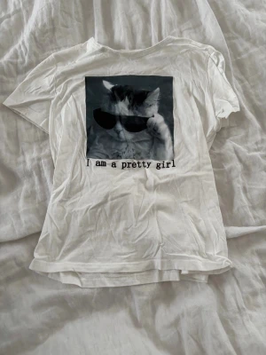 Vit t-shirt med kattmotiv - Vit t-shirt med korta ärmar och tryck framtill av en katt med solglasögon och texten 'I am a pretty girl'. Skön och enkel modell i mjukt bomullsmaterial, perfekt för dig som gillar roliga prints.🐱✨💓