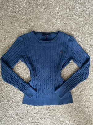 Blå kabelstickad tröja från GANT - Snygg blå kabelstickad tröja från GANT med rund halsringning och broderad logotyp på bröstet. Tröjan har långa ärmar och ribbade muddar vid ärmslut och nederkant. 