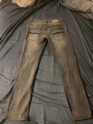 Gråa jeans från Nudie Jeans - Säljer ett par gråa jeans från Nudie Jeans med klassisk femficksmodell och raka ben. Jeansen har snygga slitningar och Nudie Jeans signaturbroderi på bakfickorna. Perfekta för en avslappnad och trendig stil.                                         Storlek: W31 L34