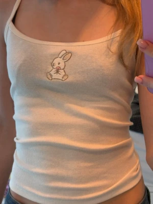 Beige ribbad linnetopp med kaninbrodyr🤍 - Supersöt vit ribbad linnetopp med smala axelband och en broderad kanin med blomma framtill. Perfekt för dig som gillar gulliga detaljer och enkel stil. Toppen är figurnära och passar till både jeans och kjol.