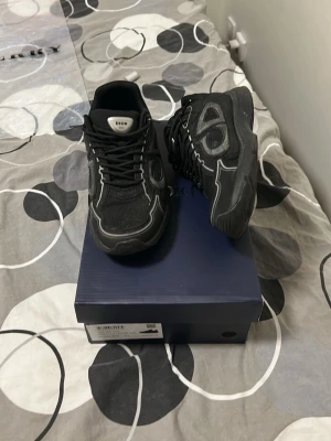 D!or B30 svarta sneakers - Säljer ett par svarta Dior B30 sneakers med vita detaljer och meshpaneler. Skorna har en chunky sula och stora Dior-loggor på sidan. Tillverkade i en mix av läder och textil för en modern look. Perfekta för dig som gillar exklusiva och trendiga sneakers.