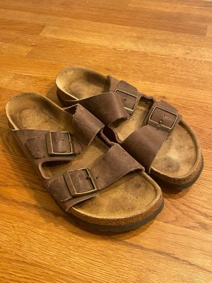 Birkenstock sandaler - Ett par klassiska bruna Birkenstock sandaler i läder med två justerbara remmar och metallspännen. De har inga defekter förutom minimala slitningar på sulan.