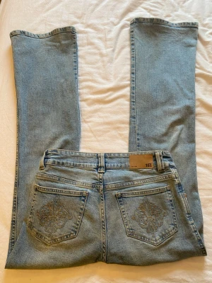 Ljusblå bootcut jeans från 157 - Säljer ett par ljusblå bootcut jeans från 157 med broderade detaljer på bakfickorna och en liten slitning vid ena knät. Jeansen har klassisk femficksmodell och är tillverkade i mjuk denim. Perfekta för en avslappnad och trendig look.