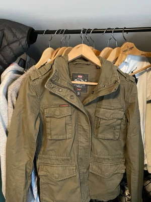 Olivgrön vindjacka från Superdry - Säljer en olivgrön vindjacka från Superdry med fyra stora fickor framtill och huva. Jackan har dragkedja och knappar en knapp är borta , samt en cool militärinspirerad look. Materialet är slitstarkt och jackan är perfekt för blåsiga dagar.