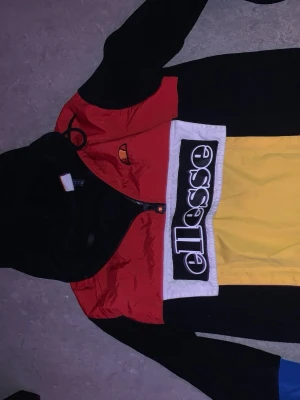 Färgglad vindjacka från Ellesse - Säljer en snygg vindjacka från Ellesse med färgblock i svart, rött, gult och blått. Stor logga framtill och dragkedja. Jackan har huva och är tillverkad i lätt, vindtätt material. Perfekt för dig som gillar streetwear och sportig stil.