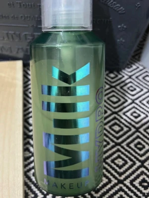 Milk Makeup Hydro Face Mist - Grön transparent sprayflaska med holografisk text och pumpfunktion. Milk Makeup Hydro Face Mist är perfekt för att återfukta huden och ge en fräsch känsla. Flaskan är cylindrisk och tillverkad i plast, med tydlig logga och modern design.