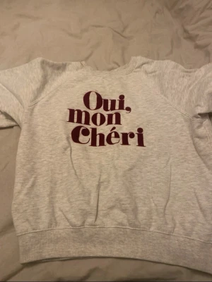 Grå sweatshirt med texttryck - Säljer en ljusgrå sweatshirt med trycket 'Oui, mon Chéri' i vinrött framtill. Tröjan har rund halsringning, långa ärmar och ribbade muddar vid ärmslut och nederkant. Perfekt för en avslappnad och trendig look.