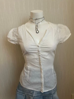 Vit kortärmad blus med knappar - Söt vit blus med korta puffärmar och dekorativa knappar framtill. Blusen har v-ringning och är sydd i ett tunt, prickigt bomullstyg som ger en romantisk känsla. Figurnära passform och lätt rynkade detaljer vid bysten för extra stil.