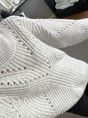 Vit stickad tröja med hålmönster - Säljer en vit stickad tröja med höghals och dekorativa hålmönster. Tröjan har en avslappnad passform, ribbstickade muddar och är gjord i ett mjukt garn som känns skönt mot huden. Perfekt för kyliga dagar och enkel att styla till olika outfits. Tröjan är från cubus och är i storlek S.