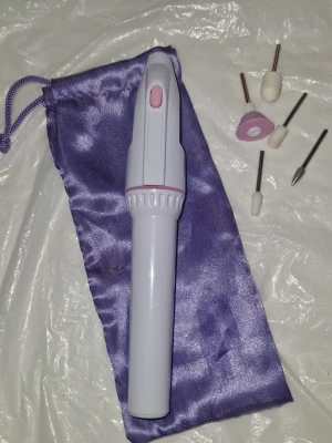 Elektrisk nagelfil med tillbehör - Elektrisk nagelfil i ljuslila med rosa knapp, levereras med fem olika sliphuvuden för filning och polering av naglar. Kommer med en snygg lila förvaringspåse i satin. Perfekt för att fixa naglarna hemma med olika former och slipytor. Drivs av ett AA batteri-medföljer.