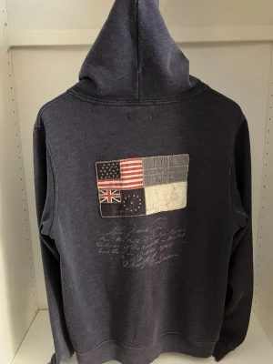 Mörkblå hoodie med flaggpatch - Mörkblå hoodie med stor patch på ryggen som visar fyra olika flaggor, inklusive USA och Storbritannien, samt broderad text under. Hoodien har huva och är tillverkad i mjukt bomullsmaterial. Perfekt för dig som gillar unika detaljer och streetwear-vibe.