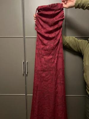 Vinröd långklänning med blommönster - Säljer en vinröd långklänning med subtilt blommönster. Klänningen är axelbandslös och har en elegant, rak siluett. Perfekt för dig som vill sticka ut med en klassisk och stilren look.