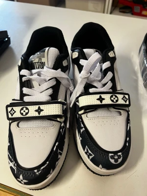 Louis Vuitton svartvita sneakers - Snygga svartvita Louis Vuitton sneakers med klassiska monogramdetaljer och bred kardborre över vristen. Skorna har vit sula med tydlig Vuitton-logga undertill och är tillverkade i läder och textil. Perfekta för dig som vill sticka ut med exklusiv streetstyle.