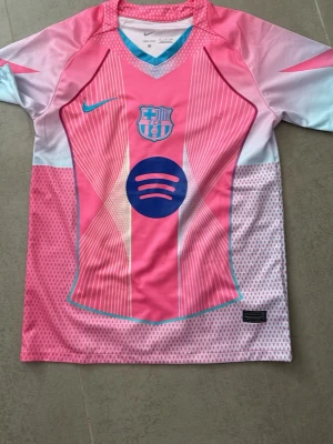 Barça matchtröja rosa Nike - Snygg FC Barcelona matchtröja i rosa och vitt med blå detaljer. Tröjan har klubbmärke och sponsorlogga på bröstet samt Nike-logga. Tillverkad i lätt och ventilerande material, perfekt för träning eller match. V-ringad hals med turkosa detaljer.